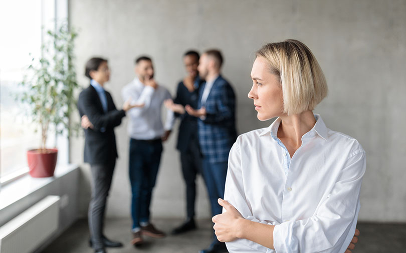 Woman standing alone while other employees gather and talk behind her.